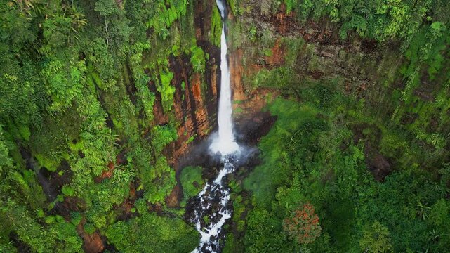 Drone view of a Kapas Biru waterfall plunging through a steep jungle canyon, framed by vibrant tropical vegetation and rugged volcanic cliffs shaping a raw and untouched natural landscape.