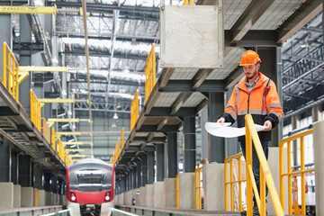 Fototapeta premium Professional Caucasian engineer standing on elevated platform holding blueprint while monitoring red commuter train inside spacious industrial workshop with symmetrical leading lines.