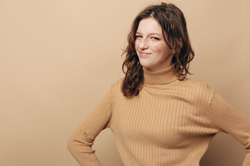 Obraz premium Smiling woman in beige turtleneck sweater poses confidently with hands on hips against plain background. Concept of casual style, confidence, and positive attitude in fashion.