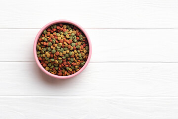 A pink bowl filled colorful dry pet food kibble on a white wooden surface. Top view. © PixelArt_25