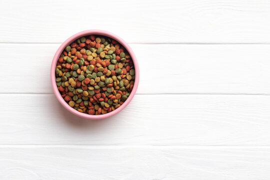 A pink bowl filled colorful dry pet food kibble on a white wooden surface. Top view.