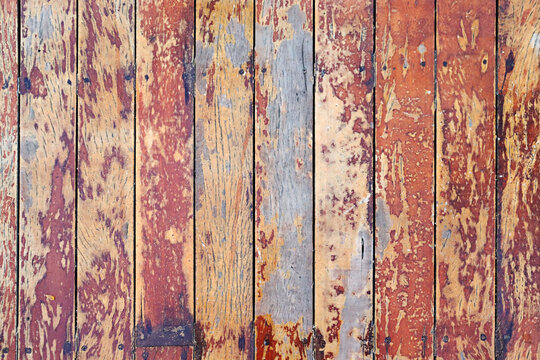A close-up view of weathered wooden floorboards with peeling paint in warm shades of red, orange, and yellow, revealing layers of aged texture beneath.