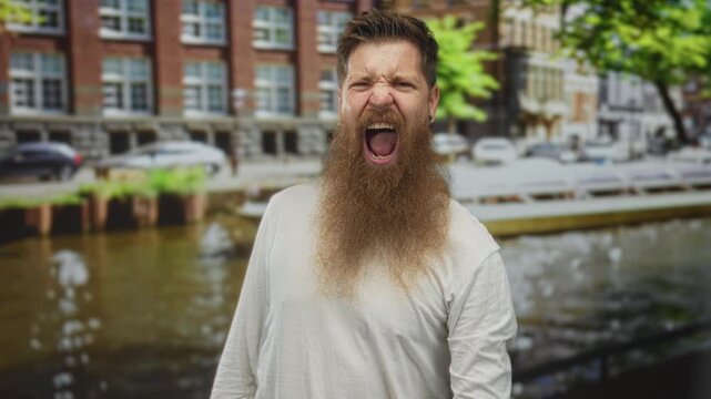Sunlit brown bearded man with tongue out making rock horns gesture on street beside canal; fun rock energy.