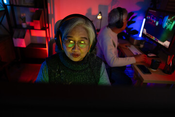 Obraz premium Senior couple, excited senior eldery Asian woman and man with headphones streamer playing video game using desktop computer at gaming room at home. colorful neon background