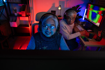 Obraz premium Senior couple, excited senior eldery Asian woman and man with headphones streamer playing video game using desktop computer at gaming room at home. colorful neon background