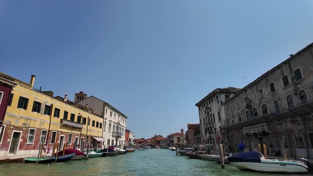 View of Venice from a boat slowly approaching across the lagoon. On either side are old houses built close to the water, boats, and footbridges. seagulls passing in the frame near the camera