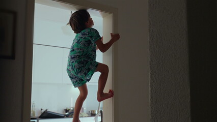Young boy scaling door frame with determined expression, gripping sides with hands and feet,...