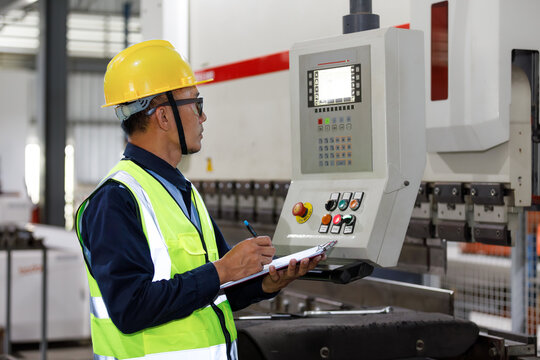 An Asian man, an industrial engineer by profession, wearing a safety helmet, operates machinery and records inspection logs in a factory.