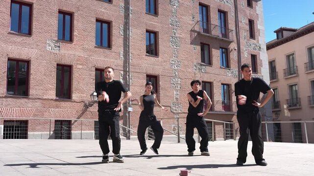 Energetic group of four contemporary dancers in black outfits performing a synchronized choreography in an urban square under bright sunlight