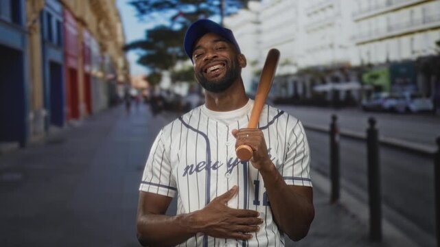 Man holding bat over shoulder on a city street wearing baseball cap and striped jersey; joy confidence.