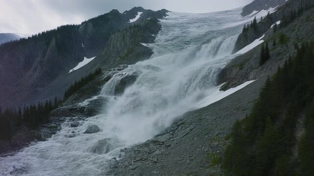A large, multi-tiered waterfall cascades down a rugged, rocky slope, surrounded by evergreen trees and patches of snow, under a cloudy sky