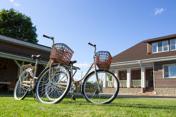 Fototapeta premium A pair of vintage bicycles with wicker baskets parked on a green lawn in front of a modern wooden house on a sunny day. Summer leisure concept with two bicycles near a country house