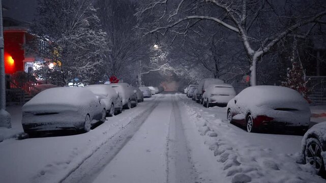Tire tracks on snow covered residential road at night. View of a city street with deep tire tracks in fresh snow and parked cars buried along the sidewalk.