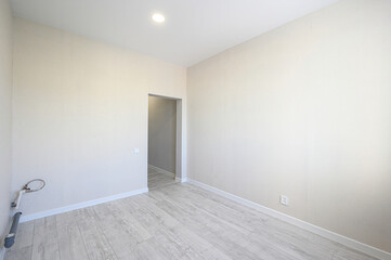 Obraz premium A spacious empty room with light beige walls, grey laminate flooring, a recessed ceiling light, a doorway, electrical outlets, and exposed plumbing pipes