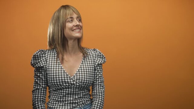 Woman smiles and gently tilts her head to the side wearing a houndstooth blouse in an orange studio; gentle joy.