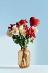 Obraz premium Beautiful bouquet of red and cream carnations in a glass vase with a red heart-shaped balloon against a light blue background with copy space
