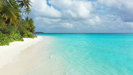 The photo shows the Maldives with crystal-clear turquoise water, soft white sandy beaches, and tall palm trees. Overwater villas stand above the calm ocean, surrounded by peaceful tropical beauty and 