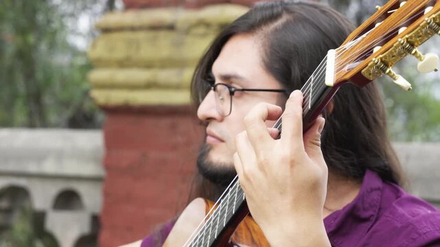 Horizontal close-up shot of a male musician with long dark hair, a goatee, and glasses. The focus is split between his serene facial expression and his left hand expertly navigating the fretboard