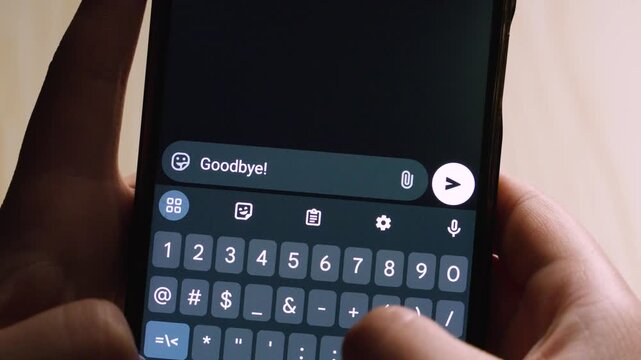 Close-up of hands holding a smartphone, typing a friendly goodbye message with a smiley face on a dark-themed digital keyboard. The predictive text bar shows options like "me," "a," and "you."
