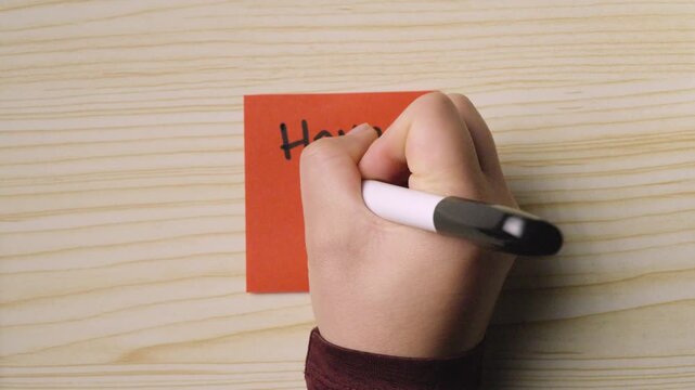 Close-up of a hand using a black marker to write "Have a Nice Day!" with a smiley face on an orange post-it note. Concept of positivity, kindness, and personal encouragement.
