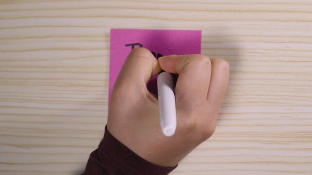 Close-up of a hand using a black marker to write "Thank You!" on a pink post-it note placed on a wooden surface. Concept of gratitude, appreciation, and personal communication.