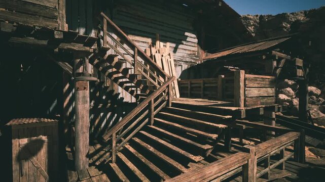 The well worn wooden stairs lead up to a rustic structure, showcasing weathered textures and shadows, surrounded by a rocky landscape under bright daylight.