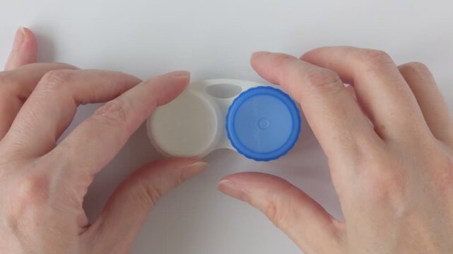 Woman's hands unscrew the lid of a contact lens case. Empty white plastic lens case with compartments for storing contact lenses. Concept for eye care and hygienic lens storage.