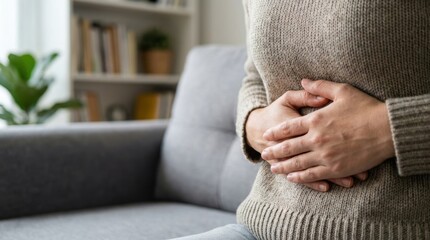 Woman's hands clasped over her abdomen indicating stomach discomfort or pain while sitting on a couch indoors
