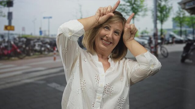 Woman smiling makes horns gesture with index fingers on crowded downtown city street; playful spontaneity.