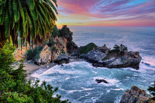 McWay Falls waterfall flowing onto a secluded beach in Big Sur, California at sunset. Turquoise Pacific Ocean waves, dramatic coastal cliffs and palm trees create a scenic Highway 1 travel destination