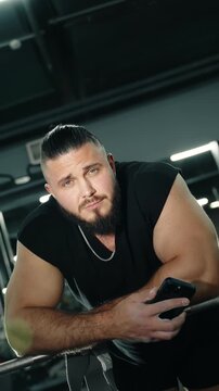 Confident bearded athlete leaning on gym equipment between sets, gripping a smartphone and meeting the camera&rsquo;s gaze under gym lighting, showcasing muscular arms and strength