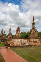 Naklejka premium Ancient ruins in Ayutthaya, Thailand. A red-brick pathway leads the viewer's eye toward a collection of diverse historical structures, including bell-shaped chedis and a tall, weathered prang.