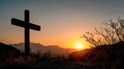 A silhouette of a cross stands on a hilltop with shrubs at sunset, casting a peaceful and spiritual atmosphere over a mountainous landscape.