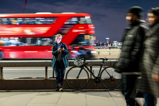 Woman standing with bicycle by London bridge at night using smartphone