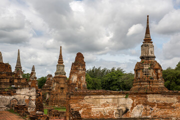Fototapeta premium Panorama of the ancient ruins in Ayutthaya's historical park under a dramatic, cloudy sky, in Thailand. Several weathered red-brick structures are visible The foreground shows the ruins of brick walls