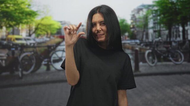 Woman pinches fingers close together showing a tiny amount with visible hand gesture on a street; playful curiosity.