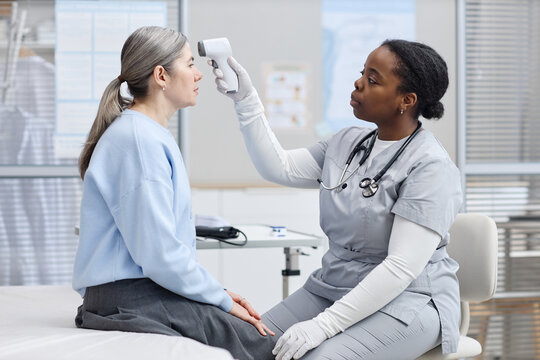 Black female healthcare worker checking temperature of middle aged Caucasian woman using digital forehead thermometer in medical clinic examination room during patient consultation