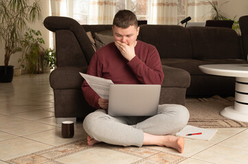 Young man sitting on floor with laptop, reading document and covering mouth in shock. Remote work...