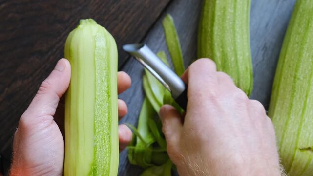 Top view first person male hands use vegetable peeler to remove zucchini skin and place peeled vegetable on table. Preparing zucchini for healthy diet. Vegetables in nutrition and vegetarian lifestyle