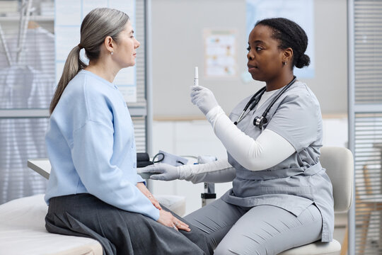 Black woman nurse examining middle aged Caucasian woman patient using medical penlight during healthcare checkup in clinic, both women sitting and facing each other in medical office