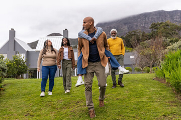 African American family walking across yard carrying teen piggyback past house with metal roofs