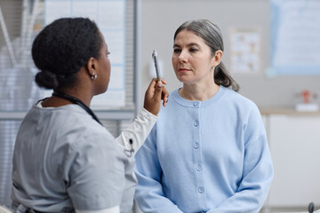 Middle aged Caucasian woman sitting while Black female doctor performing eye movement examination using pen in medical office, both women focused on assessment process © Seventyfour