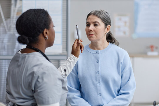 Middle aged Caucasian woman sitting while Black female doctor performing eye movement examination using pen in medical office, both women focused on assessment process