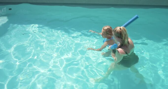 Woman holding child with noodle, armbands guiding kicking, building swim skill in backyard pool