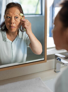 Senior Asian woman using green jade roller under eye while looking in wooden mirror at sink