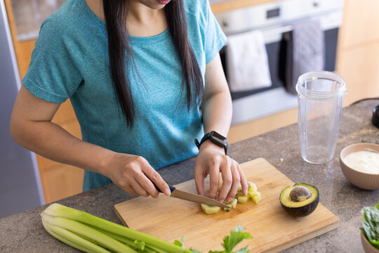Chef's knife slicing kiwi on wooden cutting board at kitchen countertop, avocado and blender nearby
