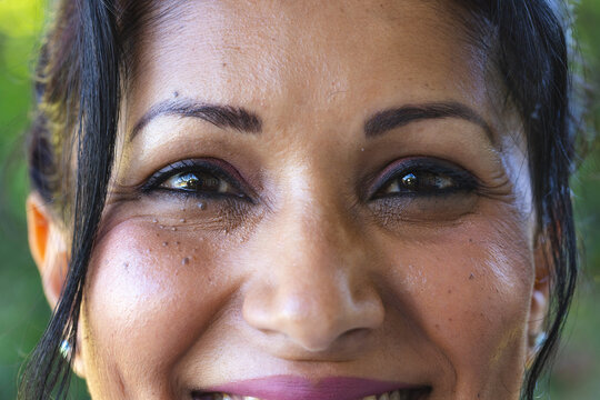 Indian woman filling frame, showing eyeliner pink lipstick and stud earring against blurred green