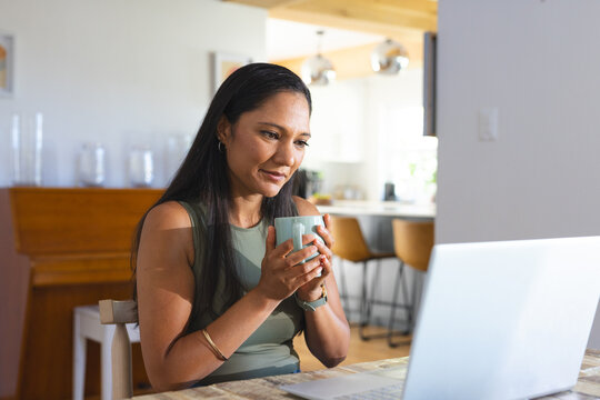 African American woman holding blue mug and gazing at silver laptop at home wearing olive top