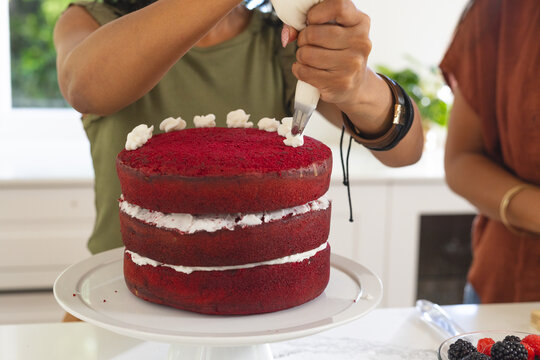 Three-layer red velvet cake receiving white rosettes from piping bag on countertop, berries nearby