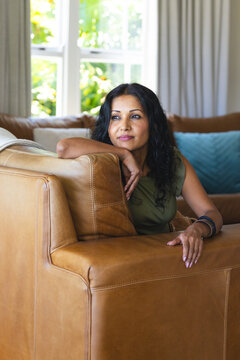 Indian woman sitting on tan leather sectional sofa, wearing olive-green top and bracelets at home